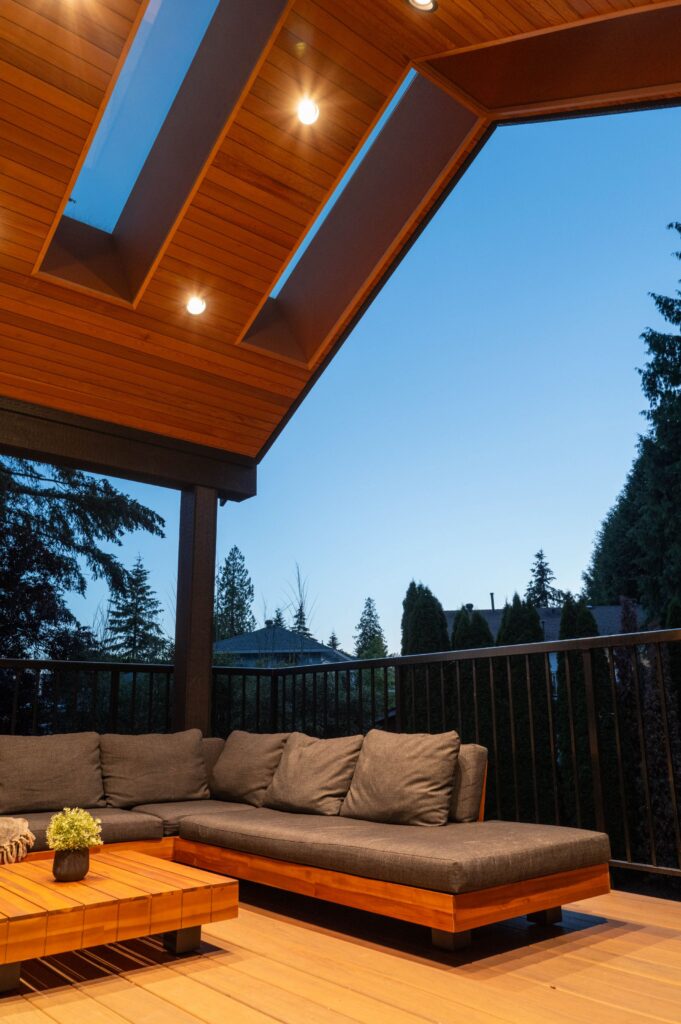 A modern outdoor patio with a wooden ceiling, skylights, recessed lights, a gray cushioned sectional sofa, a wooden coffee table with a small plant, black railings, and trees visible in the background at dusk.