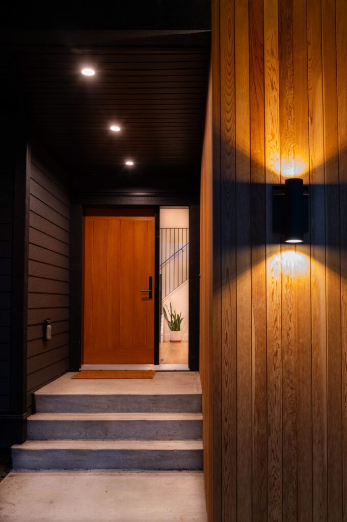 A modern wooden front door with a black handle is illuminated by wall lights. The entrance has concrete steps, vertical wood siding on the right, and dark horizontal siding on the left. Inside, a plant is visible by the stairs.