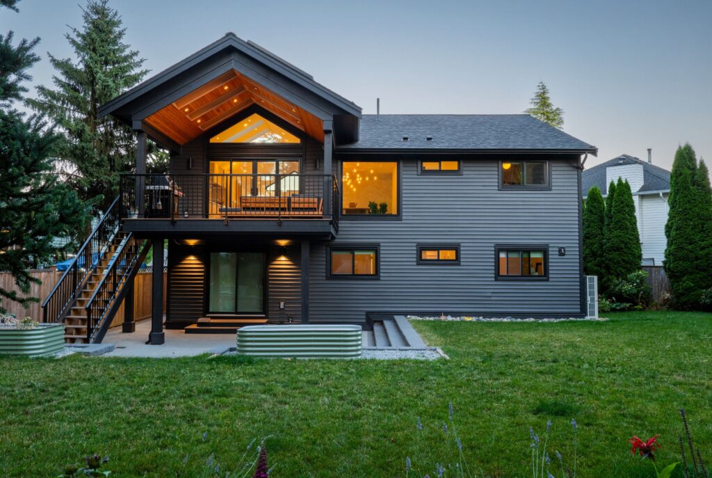 A modern two-story house with gray siding, large windows, and a covered balcony with warm lighting. The backyard features a spacious lawn, stairs, and landscaped garden beds.