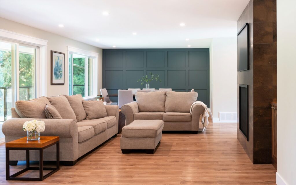 A modern living room with beige sofas, a matching ottoman, wooden floors, a dark accent wall, large windows, and a side table with flowers. Dining area is visible in the background.