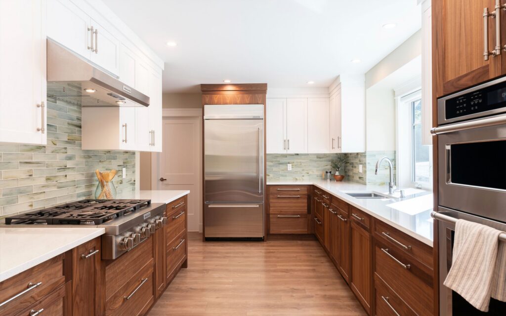 Modern kitchen with wooden lower cabinets, white upper cabinets, stainless steel appliances, including a refrigerator, oven, and gas stove, a tile backsplash, and a large window letting in natural light.