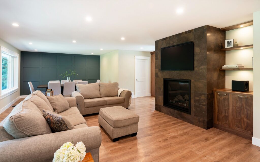 Modern living room with beige sofas, an ottoman, and wood floors. A built-in fireplace with TV above is on a stone accent wall, next to shelves and cabinets. Dining area and large window are visible in the background.