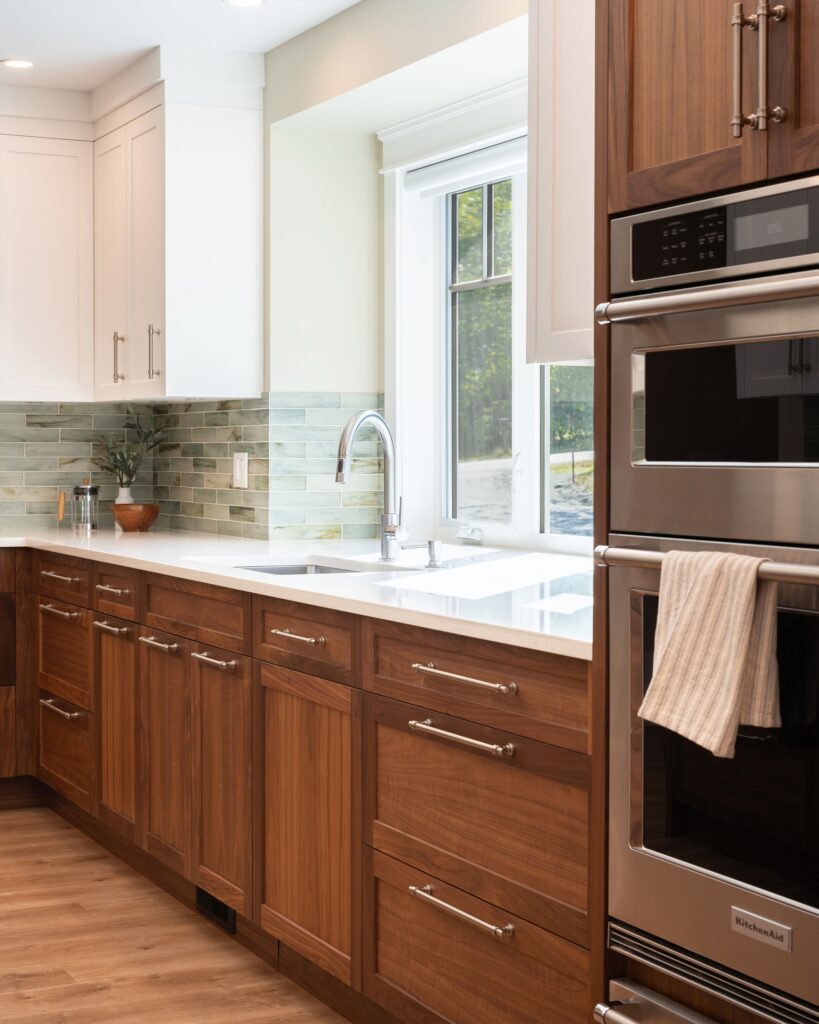 Modern kitchen with wooden cabinets, stainless steel double ovens, light countertops, a striped towel hanging from the oven handle, a sleek faucet, and a large window letting in natural light above the sink.