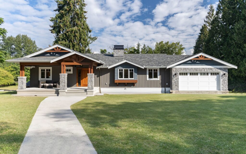 A single-story house with gray siding, white trim, a covered front porch, and attached garage, set on a spacious, well-manicured lawn with tall trees in the background under a partly cloudy sky.