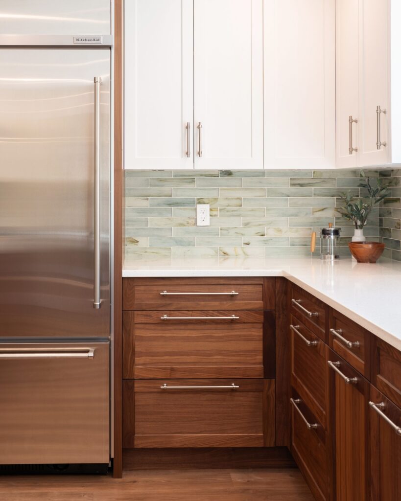 Modern kitchen with stainless steel refrigerator, white upper cabinets, wooden lower cabinets, light green tile backsplash, white countertop, and a small plant in a brown bowl on the counter.