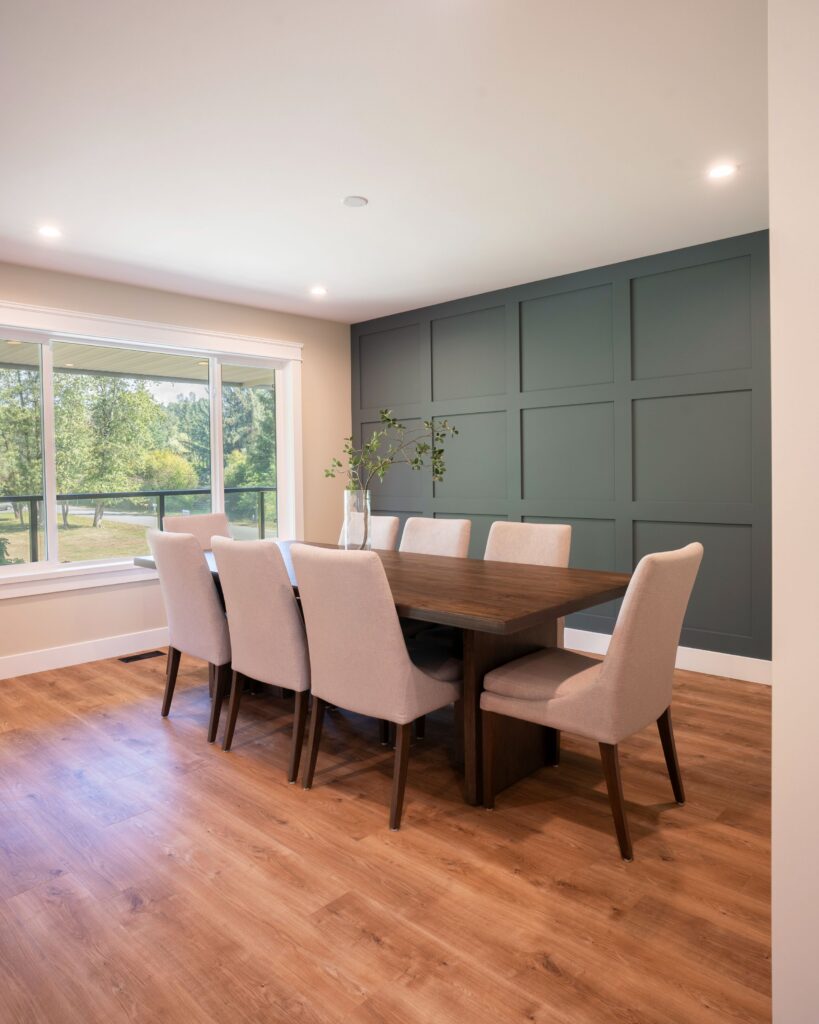 A modern dining room with a dark wooden table, eight beige upholstered chairs, a green accent wall, and a large window overlooking greenery. A vase with branches decorates the table. Light wood flooring throughout.