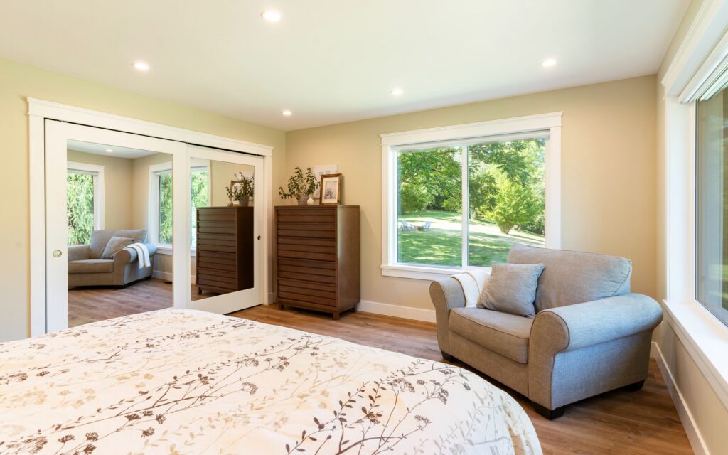 Bright bedroom with a large window, beige walls, a cozy armchair, wooden dressers, mirrored closet doors, and a bed with a patterned white and beige comforter. Green trees are visible outside.
