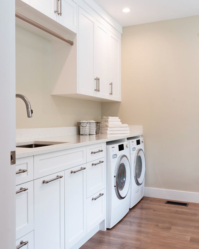 Modern laundry room with white cabinets, a countertop, stacked folded towels, a wire basket, a sink, and front-loading washer and dryer on a wooden floor under soft, recessed lighting.