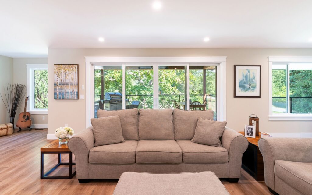 A bright living room with a beige sofa, ottoman, and side tables, large windows showing greenery outside, wall art, and a guitar in the corner on a wooden floor.