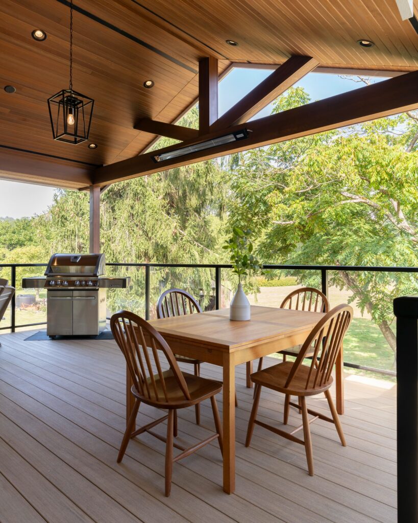 A covered outdoor patio with a wooden ceiling, a hanging lantern light, a wooden dining table with four chairs, a vase with greenery, a grill, and glass railing overlooking lush green trees.