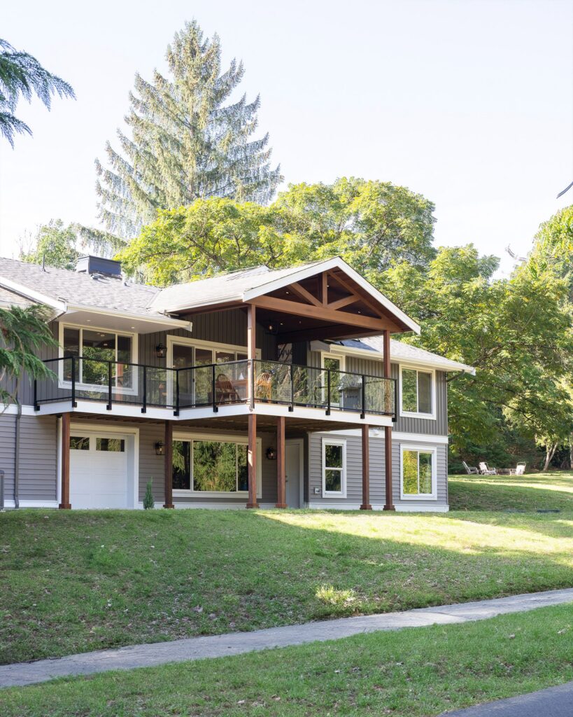 Modern two-story house with gray siding, white trim, and large windows. Features a covered balcony with glass railing, wooden beams, and a garage, surrounded by grass, trees, and a paved road.
