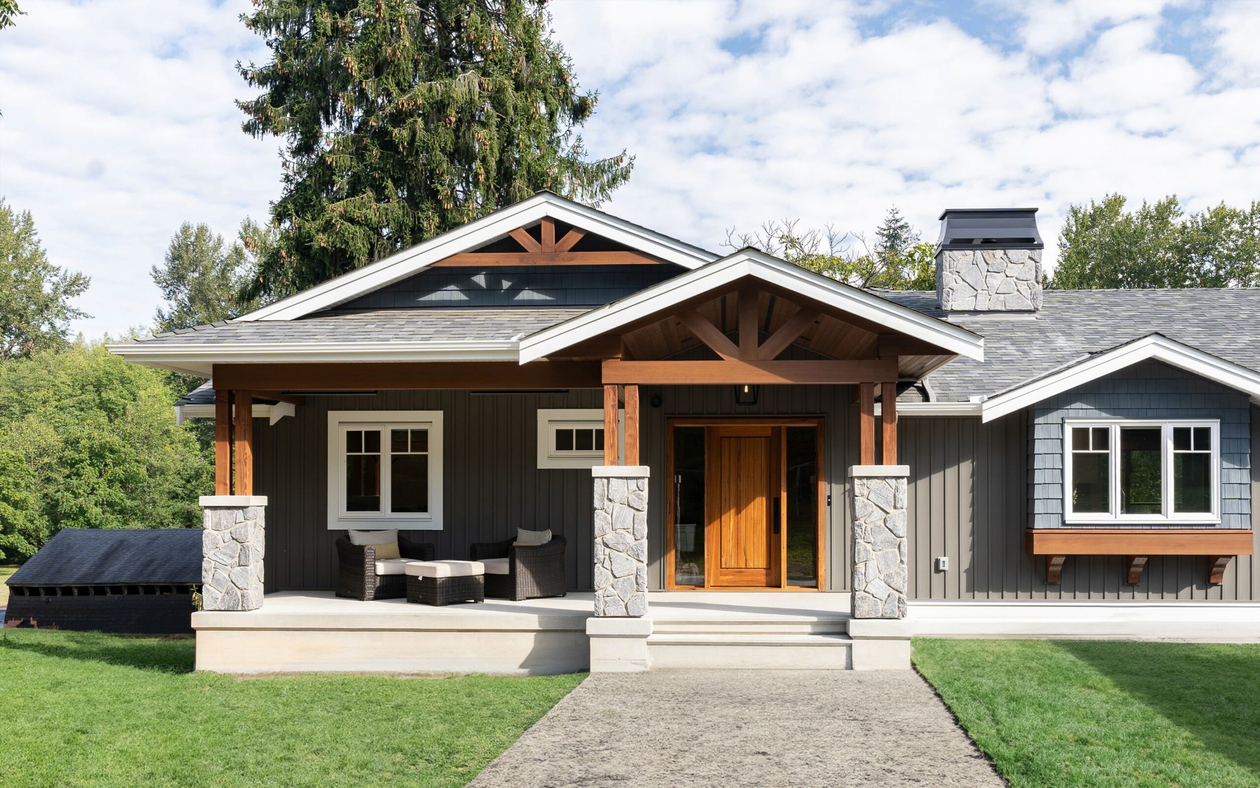A modern craftsman-style house with gray siding, white trim, a wooden front door, stone pillars, and a covered porch with outdoor seating, surrounded by a green lawn and trees.