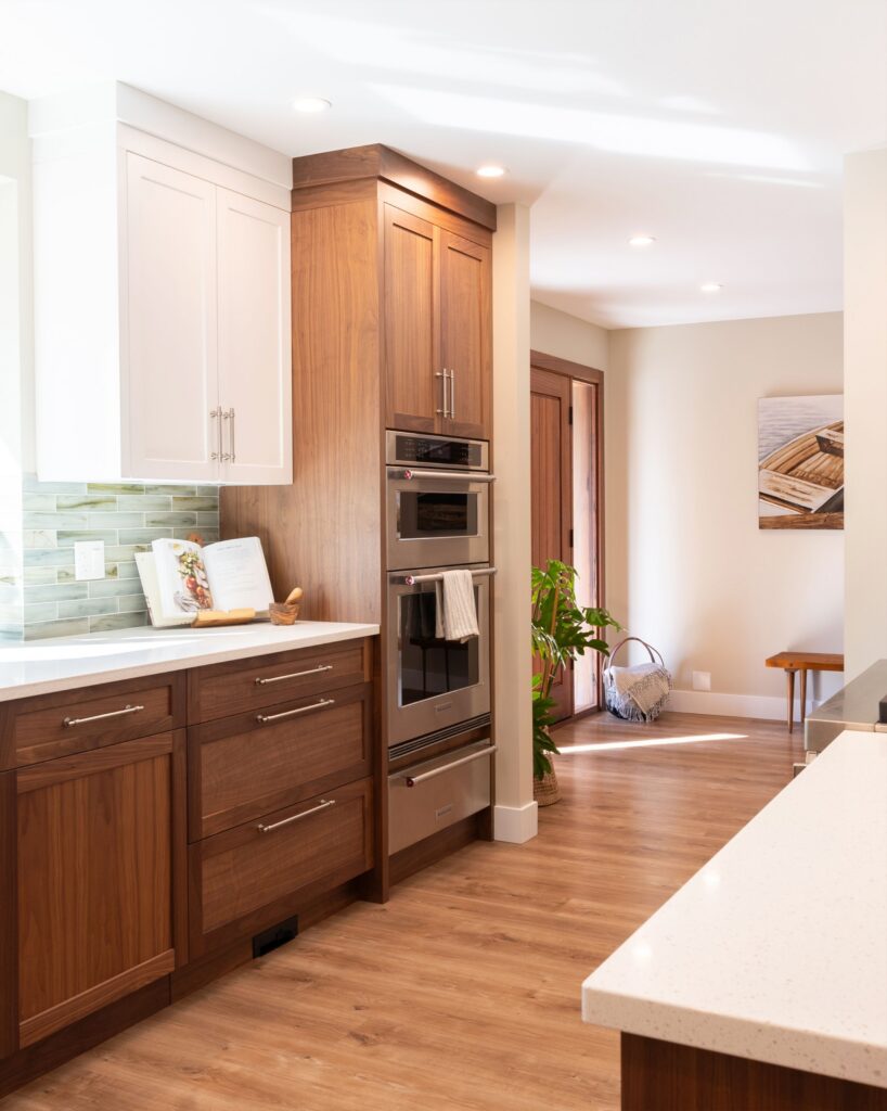 Modern kitchen with wood cabinets, white countertops, built-in double oven, and green tile backsplash. A plant and basket are near a wooden door; sunlight brightens the space with a cozy and clean atmosphere.