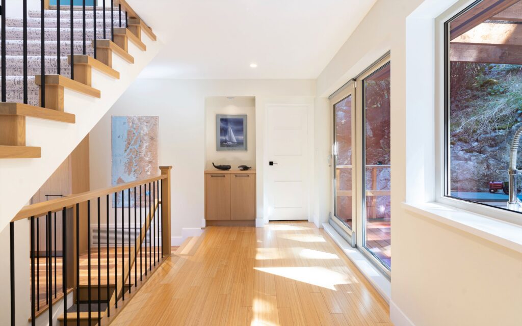 Bright hallway with light wood floors, large windows on the right, a modern staircase with black railings on the left, and a cabinet with framed artwork and decor at the end of the hall.