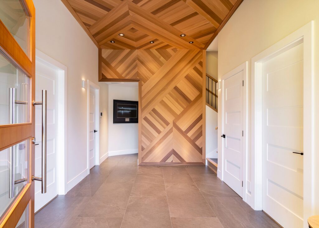 Modern hallway with a geometric wooden ceiling and accent wall, large gray floor tiles, white paneled doors, and a wall-mounted TV. Natural light enters through a glass door with a metal handle.