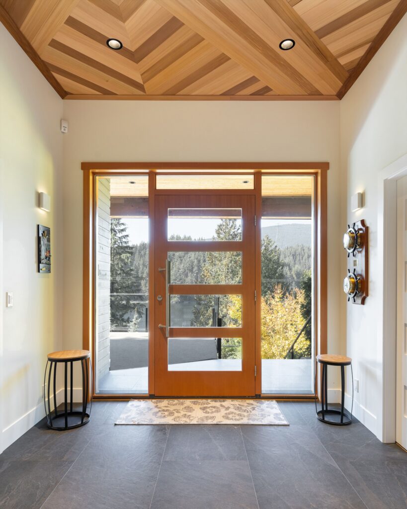 Modern entryway with large wooden door and glass panels, geometric wood ceiling, two small side tables, a wall-mounted sculpture, and windows offering a view of trees and mountains outside.
