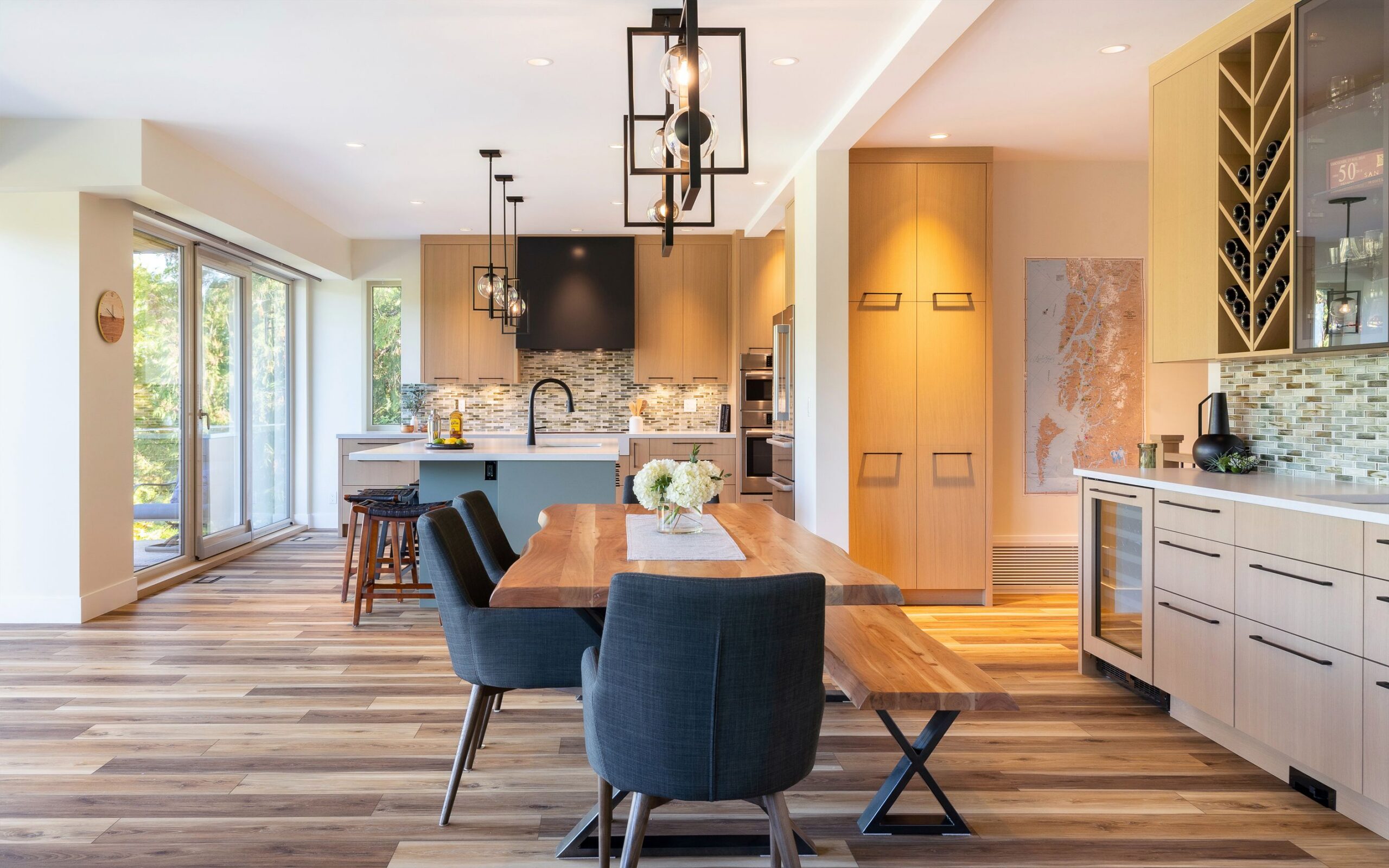 Modern open-concept kitchen and dining area with light wood cabinets, a large wooden dining table, black chairs, pendant lights, an island with stools, and large windows letting in natural light.