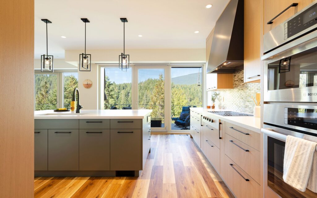 Modern kitchen with wood floors, sleek cabinetry, stainless steel double ovens, a central island with a black faucet, and large windows showcasing a scenic forest and mountain view in the background.