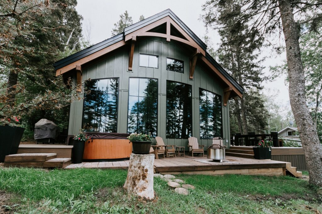A modern cabin with large windows, wooden beams, and a spacious deck featuring Adirondack chairs, potted plants, a hot tub, and surrounded by tall trees and greenery.