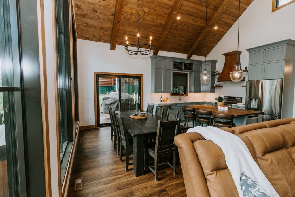 Open-concept kitchen and dining area with vaulted wood ceiling, large dark dining table with chairs, modern gray cabinets, stainless steel appliances, and natural light from large windows and sliding doors.