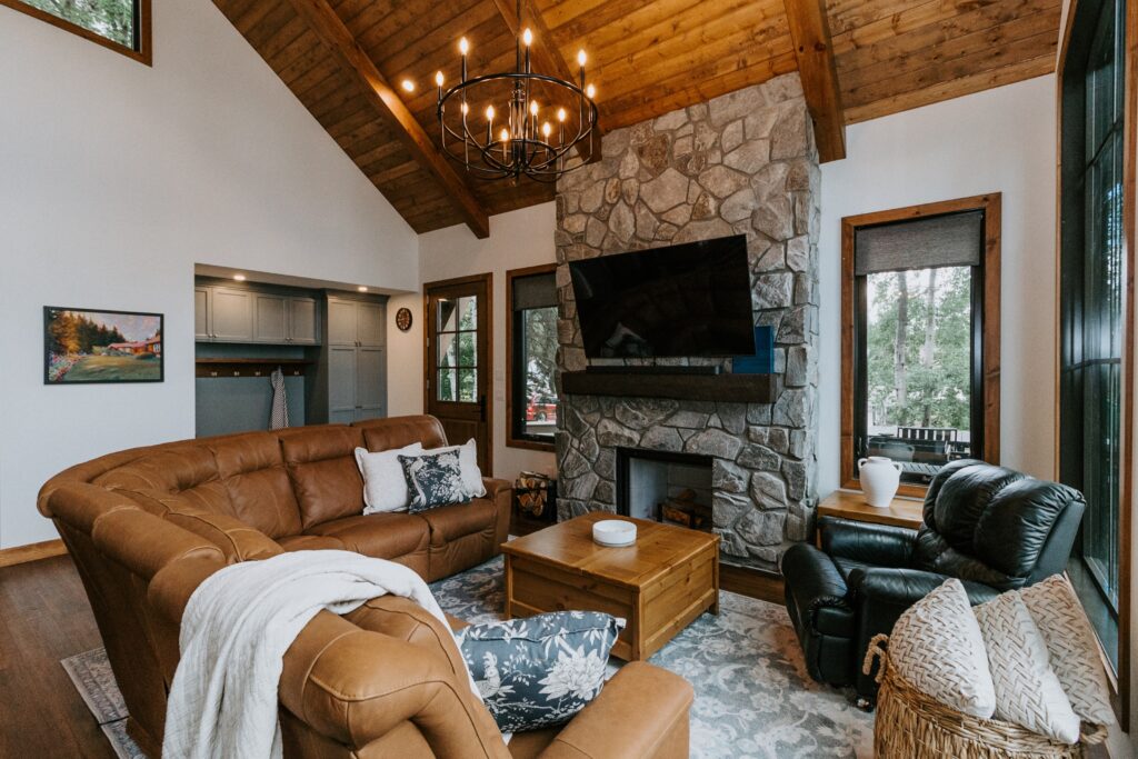 A cozy living room with a brown sectional sofa, stone fireplace, mounted TV, rustic wood ceiling, large windows, and a chandelier. There’s a coffee table, armchair, and various decorative accents. Natural light fills the space.