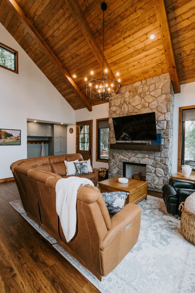 A cozy living room with a high wooden ceiling, stone fireplace, mounted TV, brown leather sectional sofa with pillows and a blanket, wooden coffee table, and large windows letting in natural light.