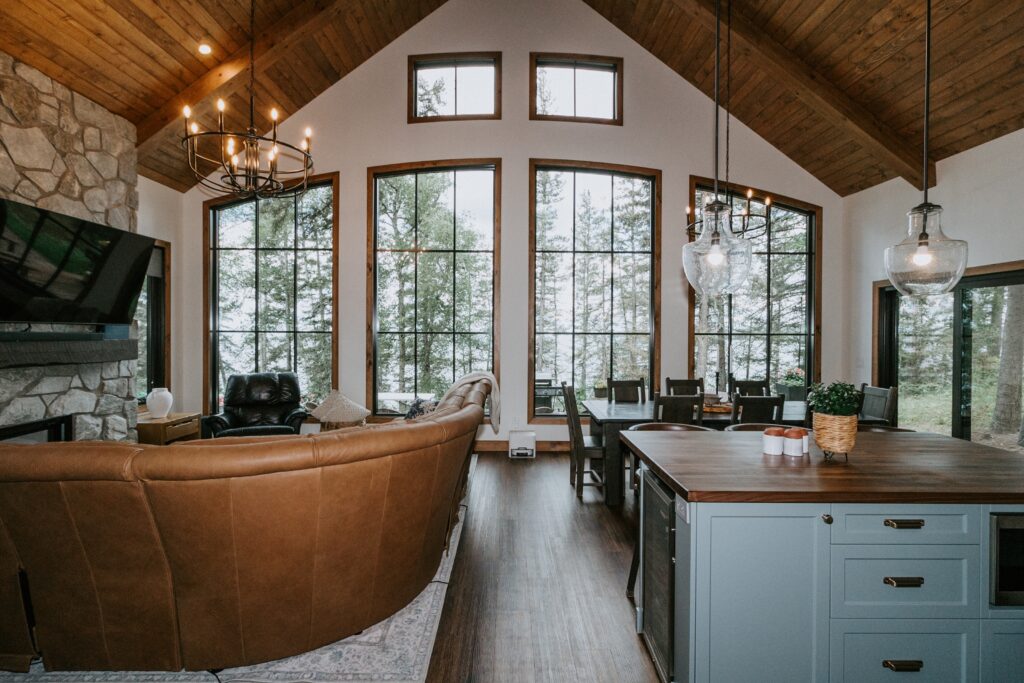 A cozy living and dining area with a brown leather sectional, stone fireplace, large floor-to-ceiling windows overlooking trees, wood ceiling, and a kitchen island with pendant lights.