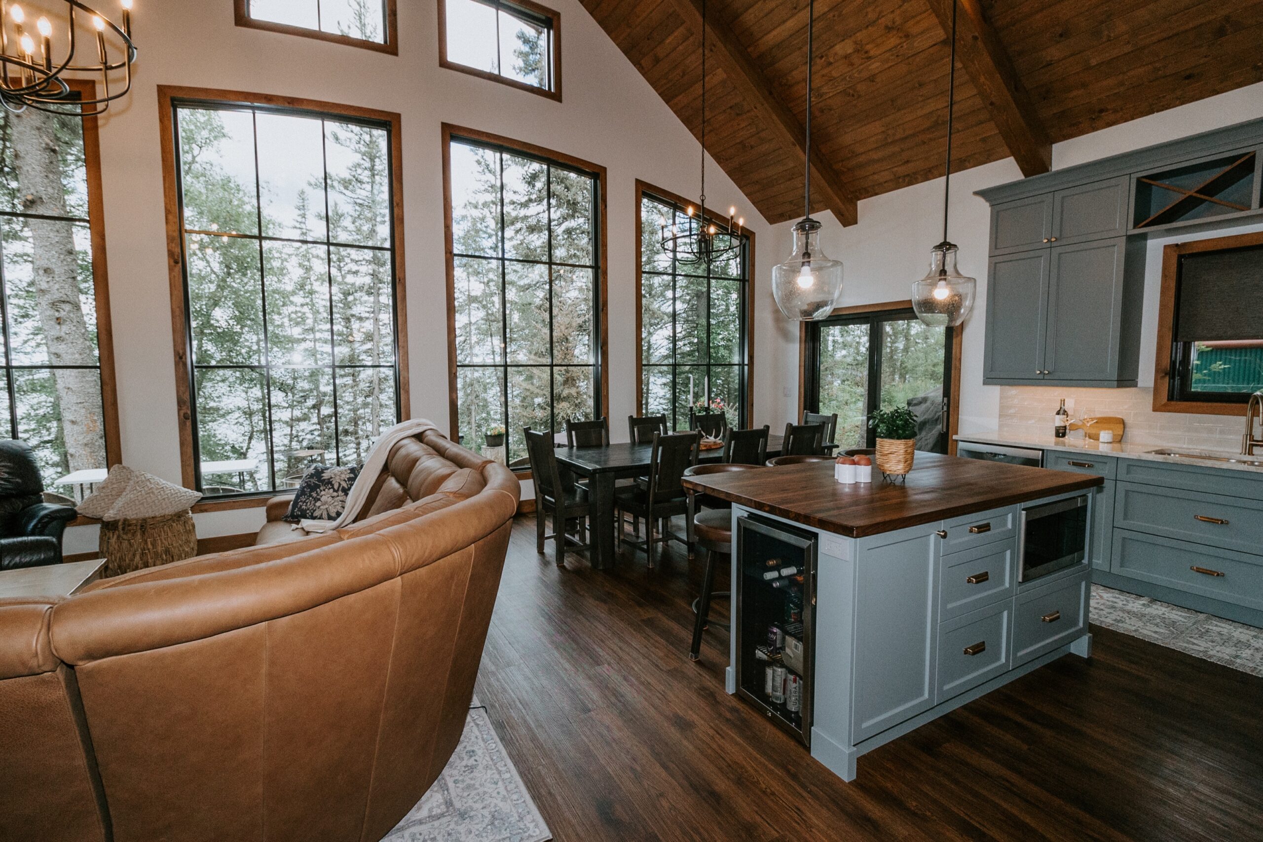 Open-concept living and dining area with large windows, vaulted wood ceiling, blue kitchen island, modern light fixtures, brown leather sofas, and a dark wood dining table. Trees and snow are visible outside.
