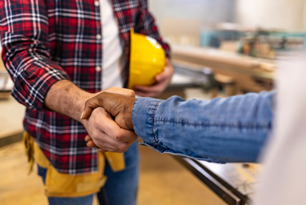 Two people shaking hands indoors, one wearing a red plaid shirt, tool belt, and holding a yellow hard hat, the other in a denim jacket. The setting appears to be a workshop or construction site.