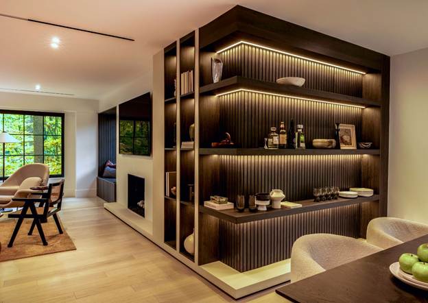 A modern, warmly lit living and dining area features built-in open shelves displaying decor, books, and bottles. The space has light wood floors, neutral furnishings, and large windows showing greenery outside.