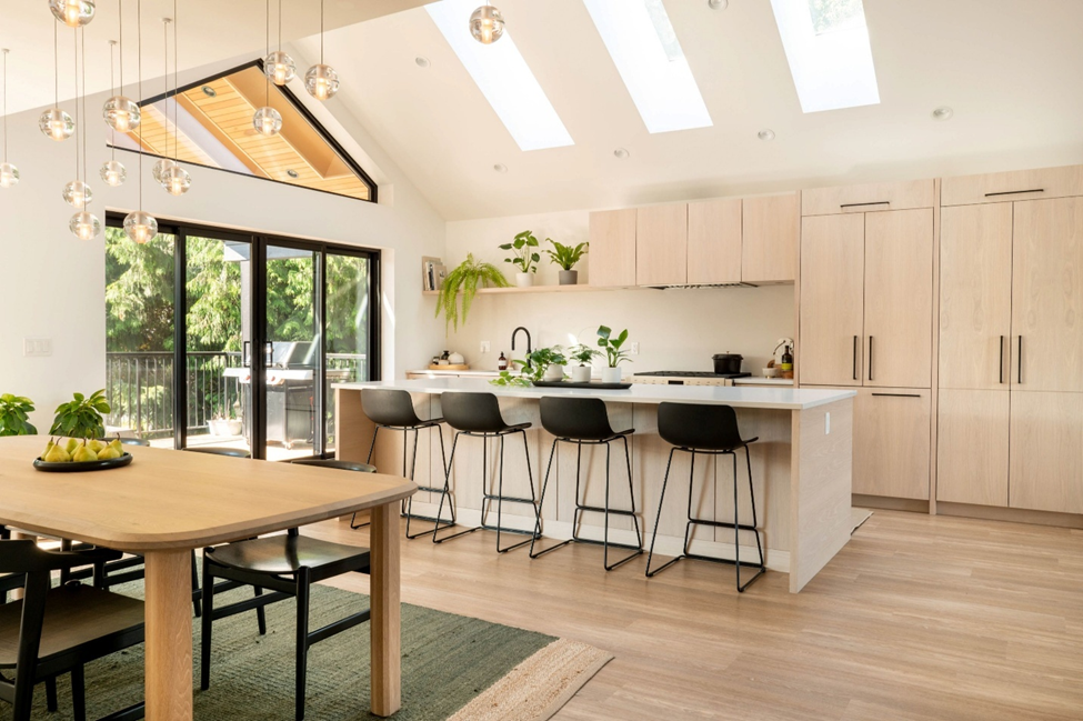Bright, modern kitchen and dining area with light wood cabinets, black barstools at a large island, skylights, hanging lights, and large windows overlooking greenery. Green plants and fruit decorate the space.