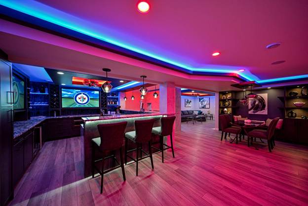 Modern basement lounge with bar, barstools, neon and recessed lighting, wall art, a TV displaying a Winnipeg Jets logo, a seating area with armchairs, and hardwood floors.