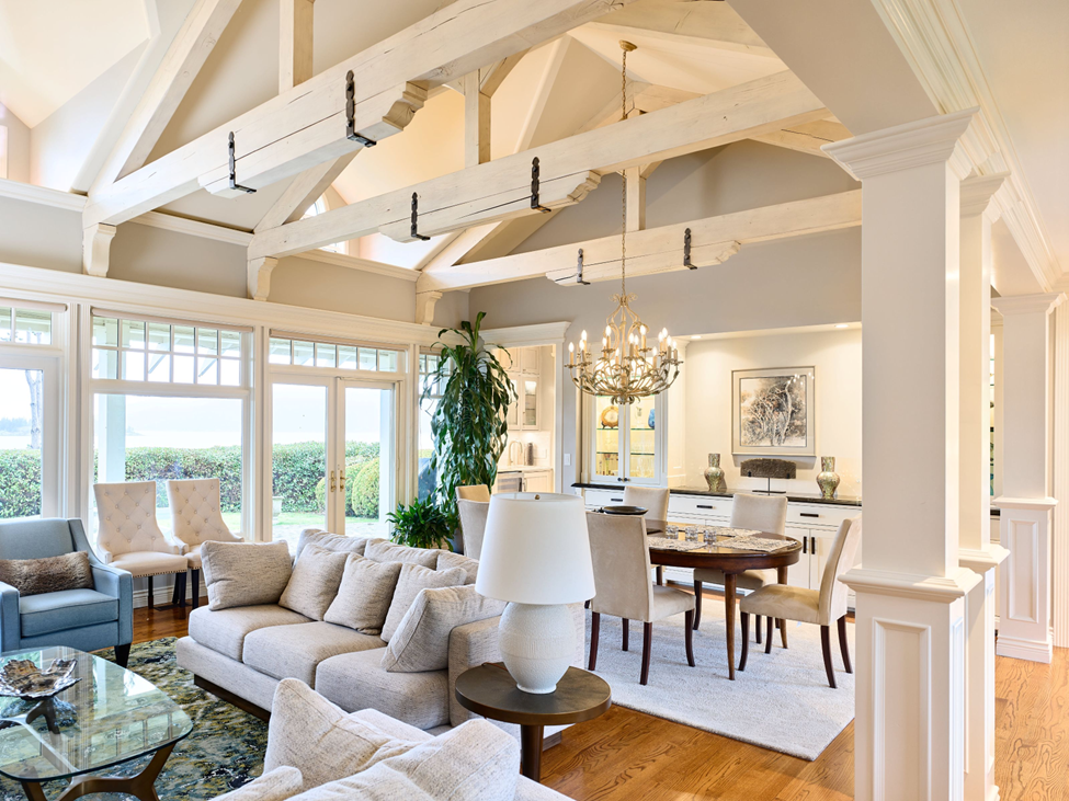 Bright, elegant living and dining room with high, beamed ceilings, large windows, neutral-colored furniture, a chandelier, and hardwood floors. Natural light fills the open space, creating a cozy, inviting atmosphere.