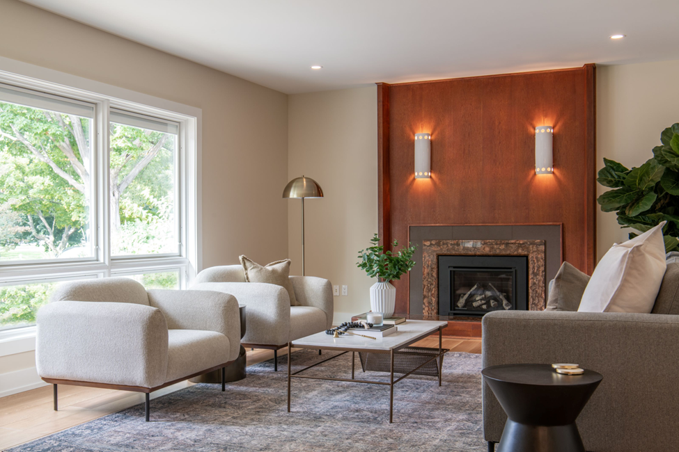 A modern living room with large windows, two light gray armchairs, a gray sofa, a coffee table with decor, a leafy plant, a floor lamp, and a fireplace set in a wooden accent wall with two sconces.