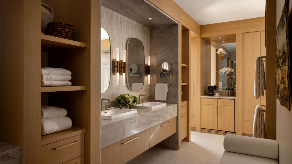 Modern bathroom with light wood cabinetry, a double sink vanity, marble countertops, oval mirrors, and wall sconces. This stylish space by RenoMark members was featured in the CHBA National Awards 2026. A small kitchenette is visible in the background.