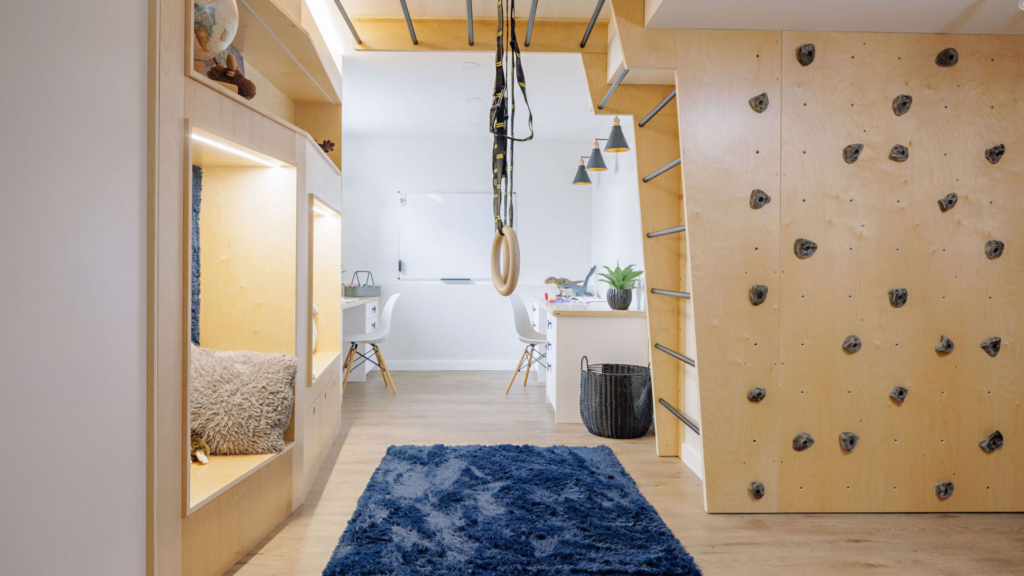 Modern room with a blue rug, cozy reading nook, wooden climbing wall, and gymnastic rings—an inspiring space reminiscent of RenoMark members CHBA National Awards 2026 style, featuring natural wood and white tones, plus a workspace with two white desks.