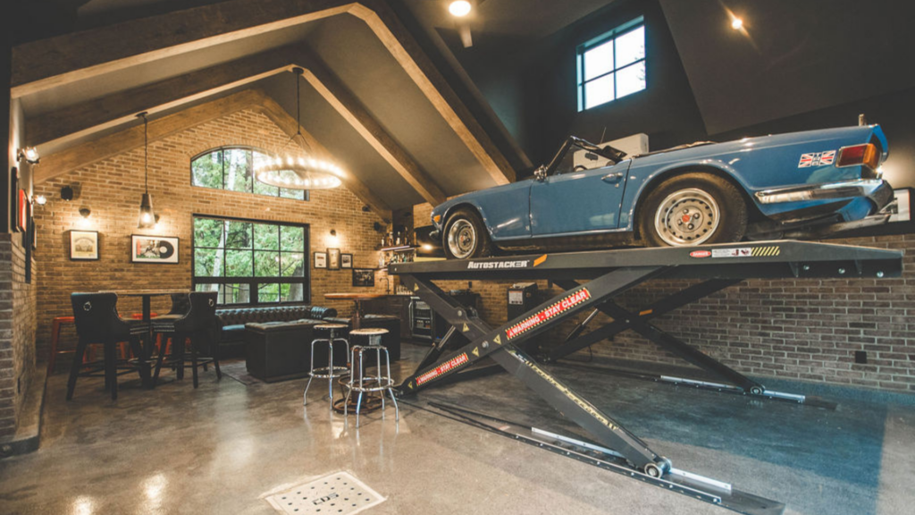 A classic blue convertible car is elevated on a hydraulic lift inside a stylish garage—worthy of RenoMark members CHBA National Awards 2026—with brick walls, modern lighting, large windows, and a chic lounge area with chairs and a table.