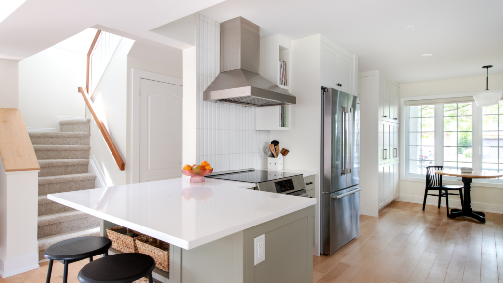 Bright modern kitchen with white cabinets, stainless steel fridge and range hood, electric stove, island with stools, and a bowl of fruit—crafted by RenoMark members CHBA National Awards 2026 finalists. Wooden floors and dining area by large windows.