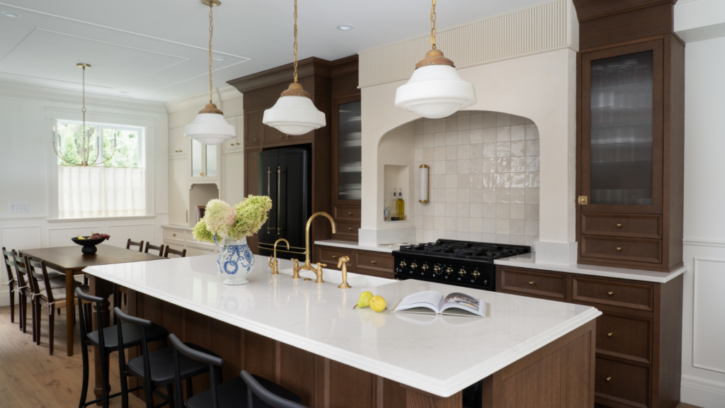 A modern kitchen by RenoMark members features a large white island, dark wood cabinets, gold fixtures, and white pendant lights. The stylish space is worthy of CHBA National Awards 2026 recognition, with a tiled backsplash and fresh lemons on the island.