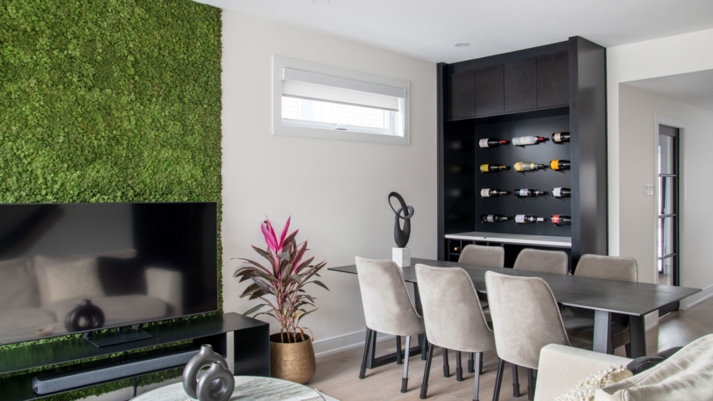 Modern dining room with a table and six chairs, wall-mounted wine rack, TV stand, potted plant, and abstract sculpture. A vibrant green moss wall glows with natural light—a space worthy of RenoMark members CHBA National Awards 2026.