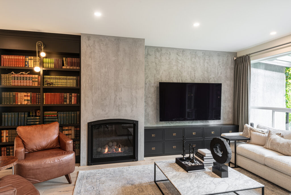 A modern living room features a leather armchair, a built-in bookshelf, a fireplace, a large flat-screen TV, a beige sectional sofa, and a coffee table with decorative items on a light rug. Large windows let in natural light.