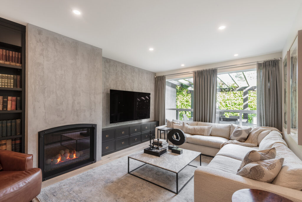 Modern living room with a light gray sectional sofa, glass coffee table, built-in fireplace, wall-mounted TV, large windows with gray curtains, bookshelves, and neutral décor. Natural light fills the space.