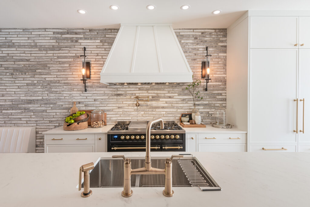 Modern kitchen with a marble island, gold faucet, farmhouse sink, black stove with brass details, white hood, and gray brick backsplash. Decorative items and lighting fixtures complete the clean, elegant look.