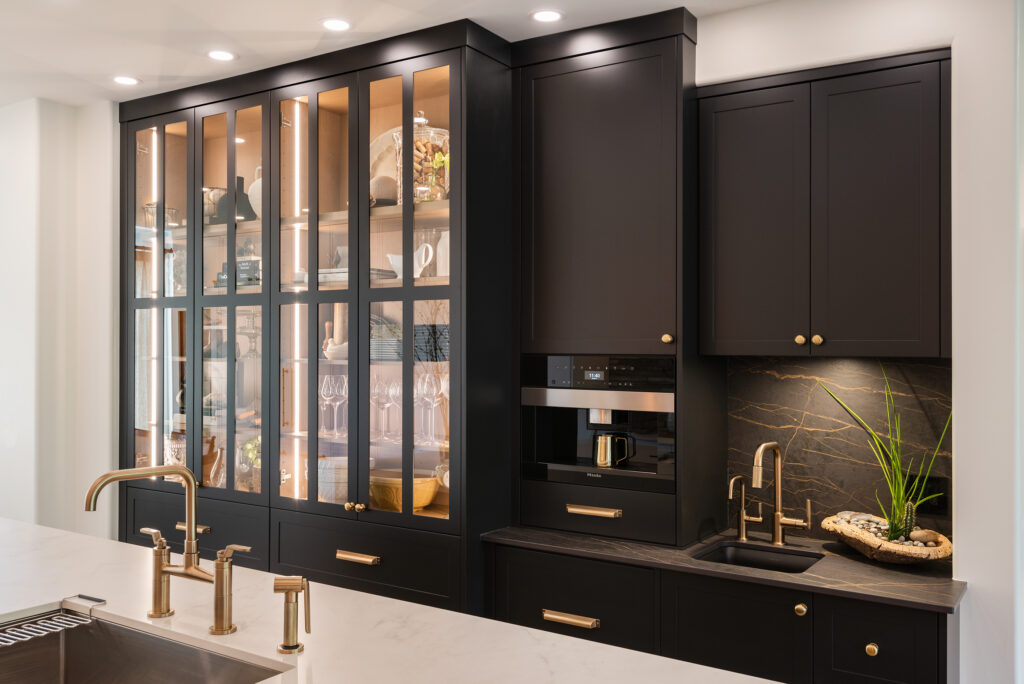 Modern kitchen with dark cabinets, gold hardware, glass-front display shelves with dishes, built-in coffee machine, small sink, marble countertop, under-cabinet lighting, and a decorative plant on the right.