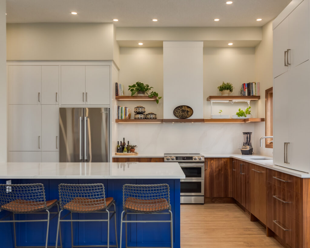 Modern kitchen with a large blue island, three metal bar stools, stainless steel fridge, wood lower cabinets, white upper cabinets, open shelves with plants and books, and recessed lighting.
