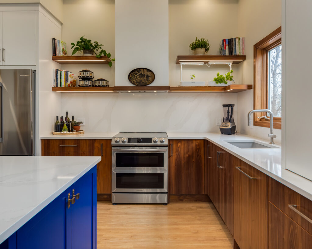 Modern kitchen with wood cabinets, white countertops, a stainless steel oven, and open shelves holding plants and books. A large window is above the sink, and a blue island is in the foreground.