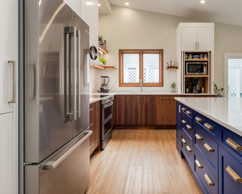 Modern kitchen with stainless steel refrigerator, wooden cabinets, blue island with gold handles, white countertops, appliances, and a window letting in natural light.