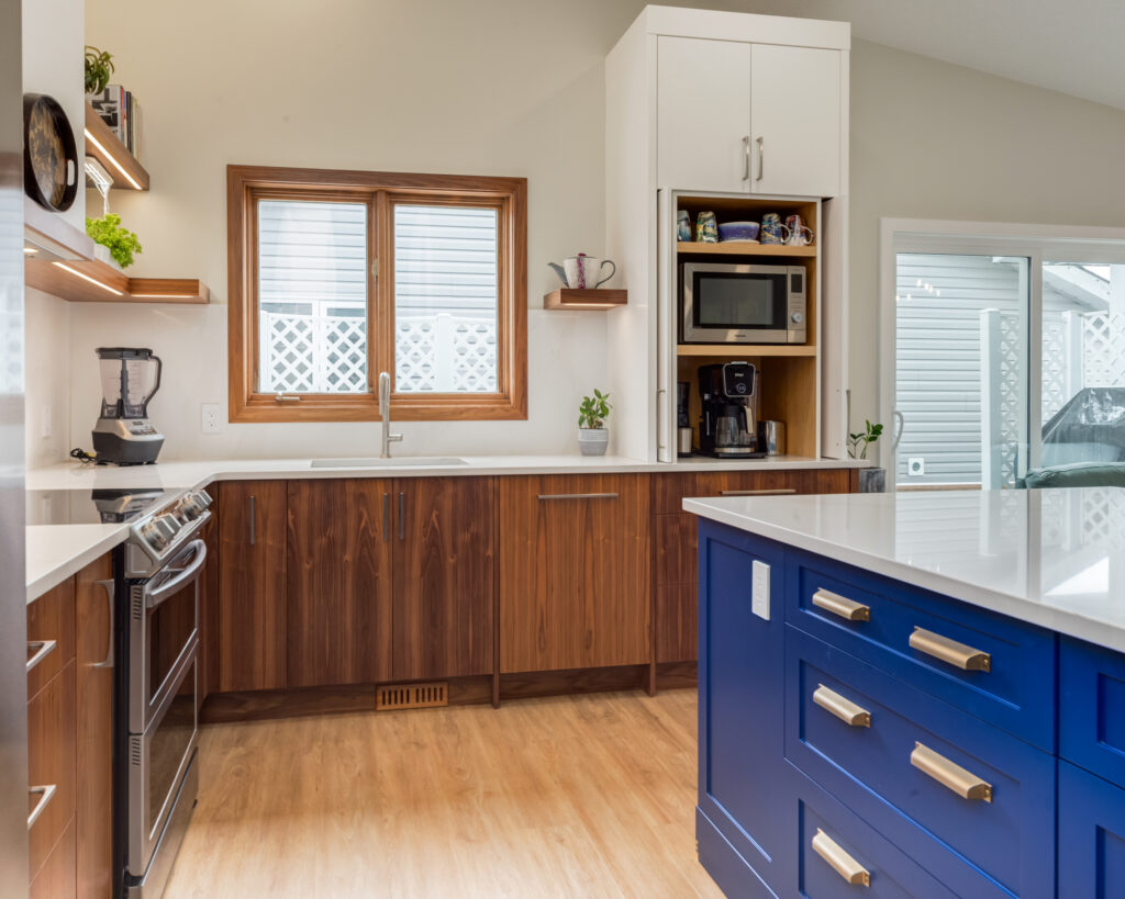 Modern kitchen with wood cabinets, white countertops, a stainless steel stove, blender, and sink under a window. There is a blue island with gold handles and open shelves with plants and mugs. A coffee station is built into the cabinetry.