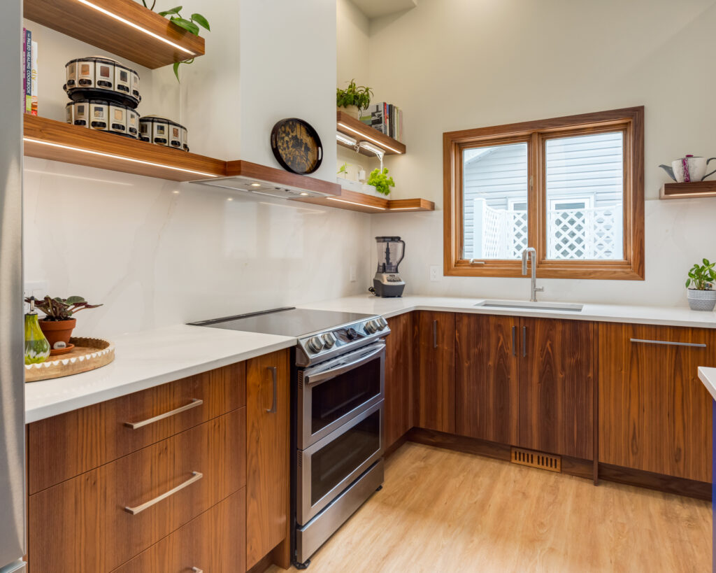 Modern kitchen with wood cabinets, white countertops, an electric stove, a double sink under a window, open shelves with plants, books, and jars, and light wood flooring. Natural light enters through the window.