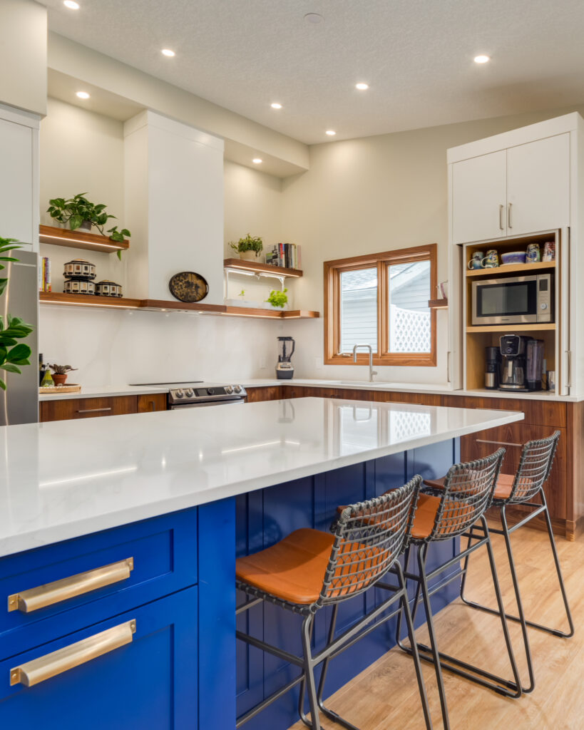 Modern kitchen with a bright blue island, three metal barstools with brown seats, white countertops, wood accents, open shelving, built-in appliances, and under-cabinet lighting. A window lets in natural light.